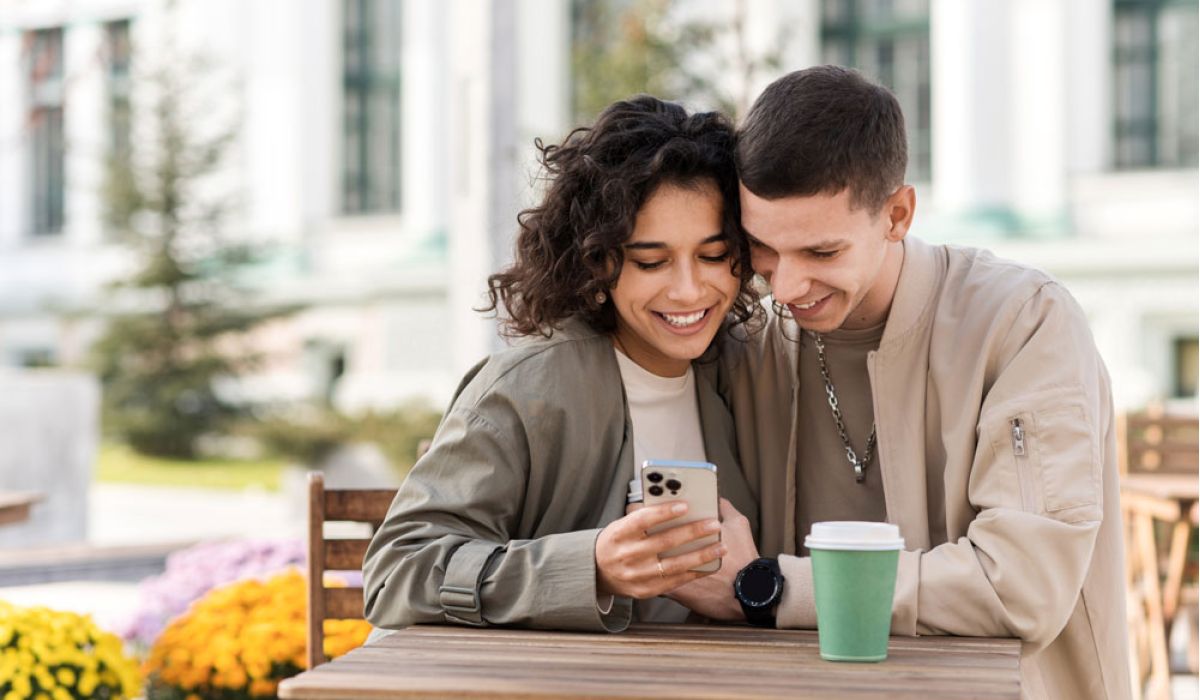 A man and woman are seated at a table, sharing a cup of coffee and looking at a cell phone together.